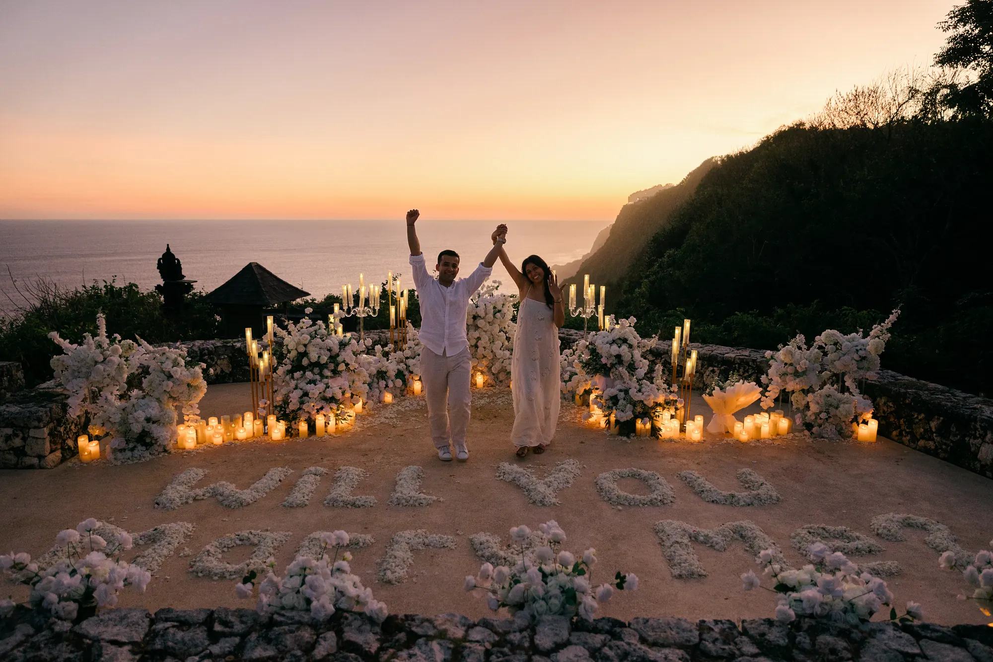 Couple celebrating with arms raised at their clifftop proposal setup at sunset in Bali