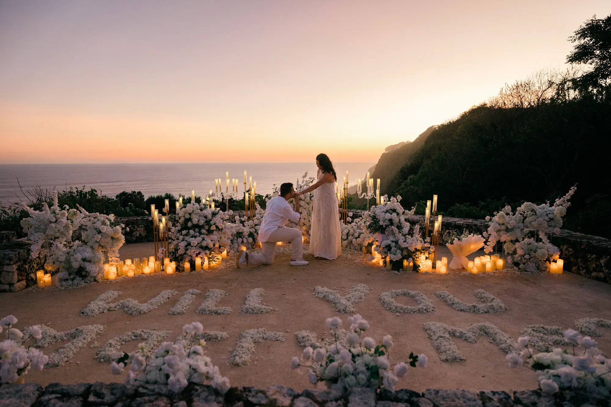Cliff proposal in Bali at sunset with 'Will You Marry Me' spelled in petals above the ocean