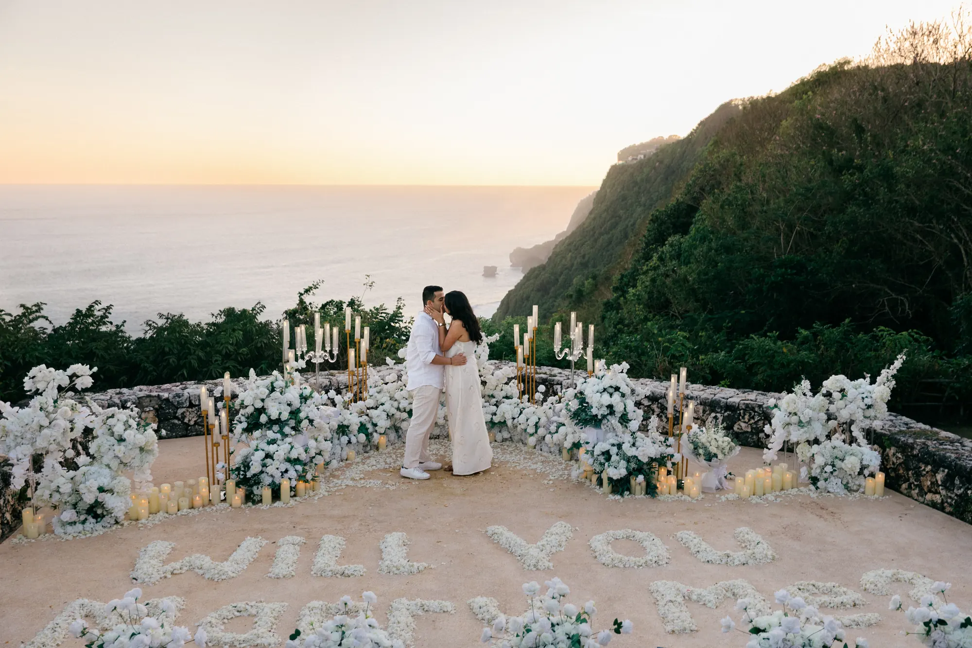 Wide view of a clifftop proposal setup in Bali with 'Will You Marry Me' in petals at sunset