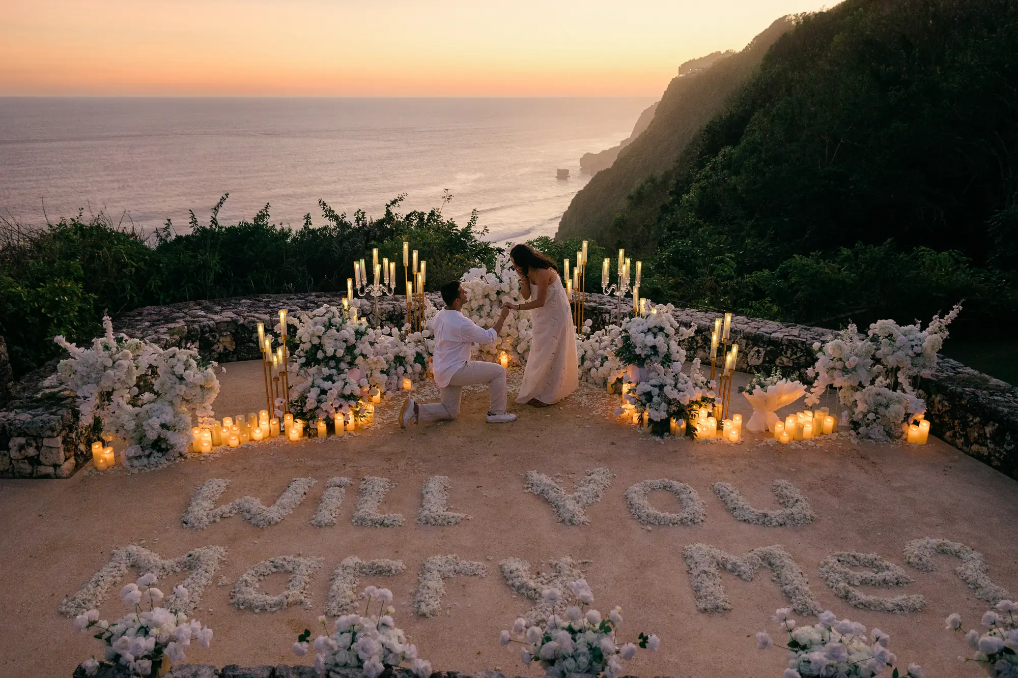 Candlelit clifftop proposal setup in Bali with white flowers and 'Will You Marry Me' at sunset