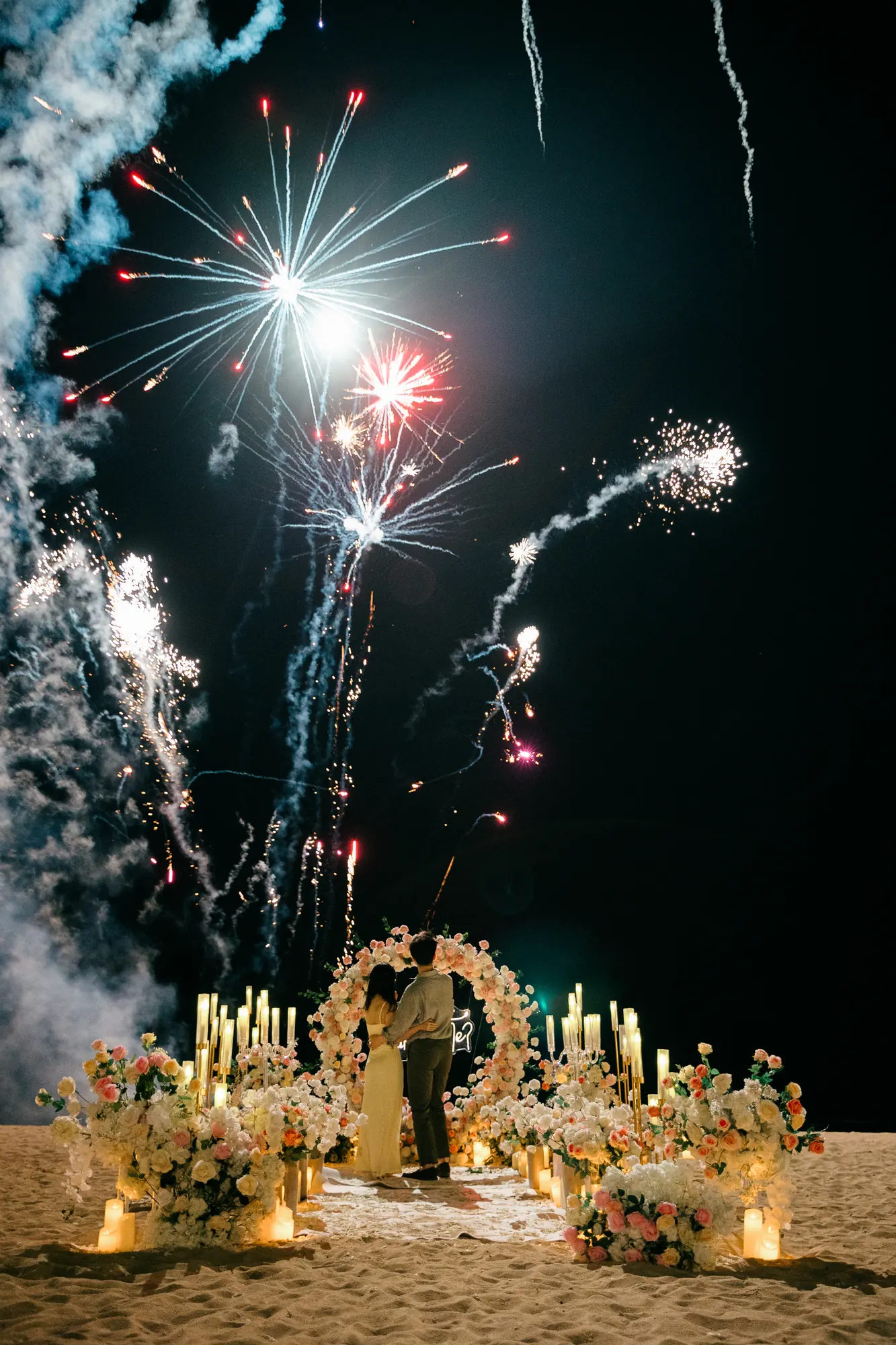 Fireworks over a candlelit floral arch as a man kneels to propose on a Bali beach