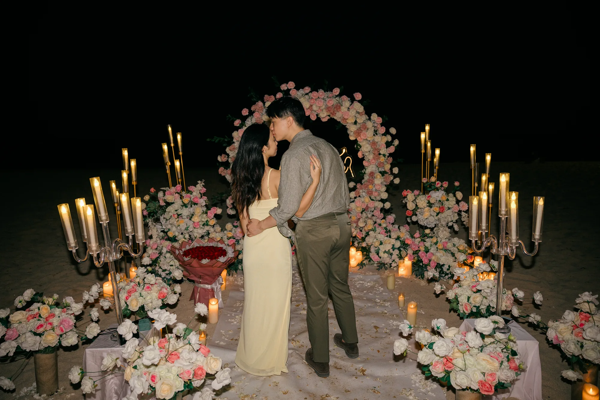 Couple embracing at a candlelit floral arch on the beach at night in Bali