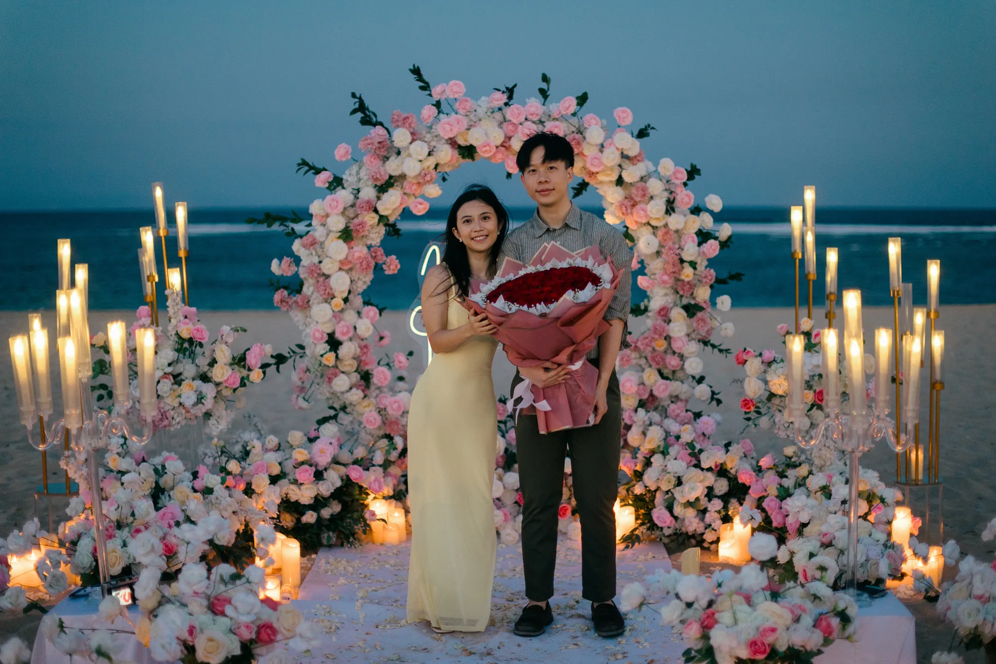 Couple holding red roses at a candlelit floral arch on a Bali beach at night
