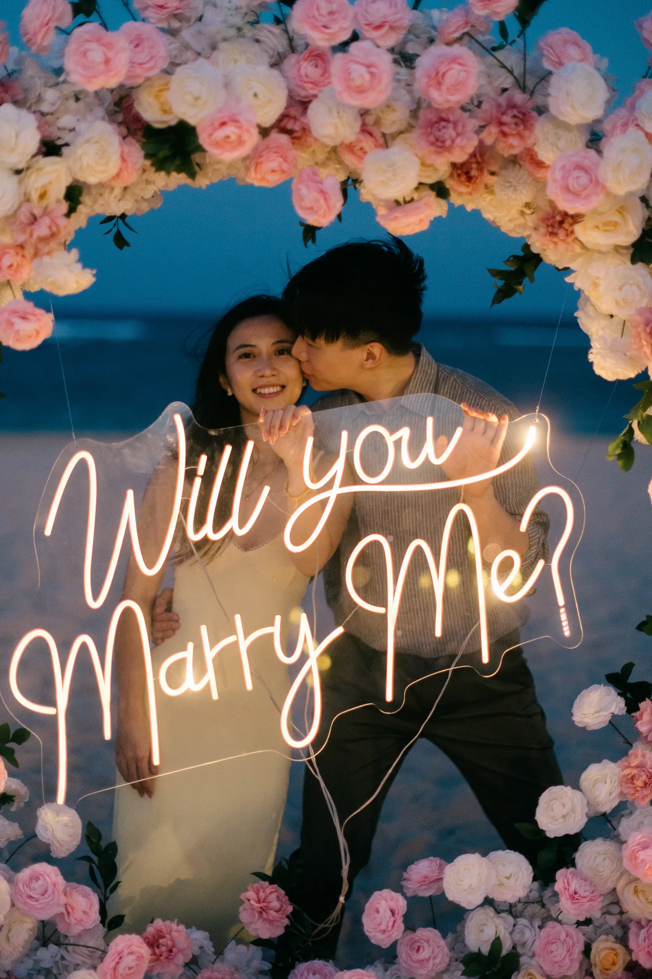 Couple embracing under a floral arch with a 'Will You Marry Me' neon sign in Bali