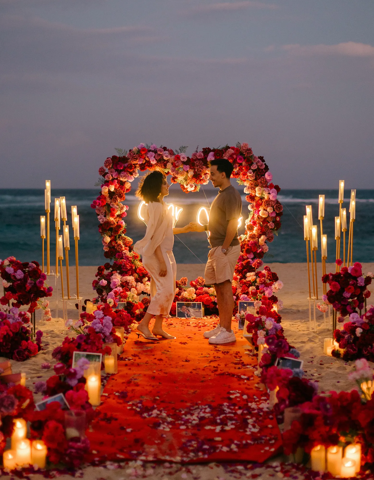 Couple at a glowing red rose heart arch at dusk on a Bali beach