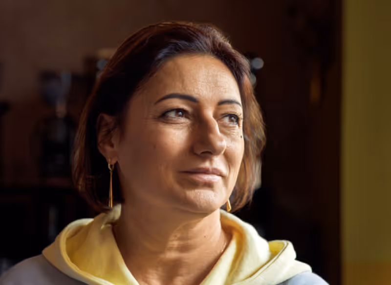 Portrait of a woman with short brown hair wearing gold earrings and a yellow hoodie looking to the side.