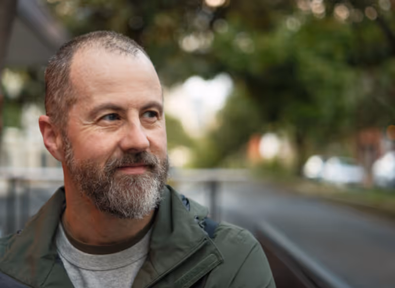 Bearded man in a green jacket looking to the side with a blurred outdoor background.