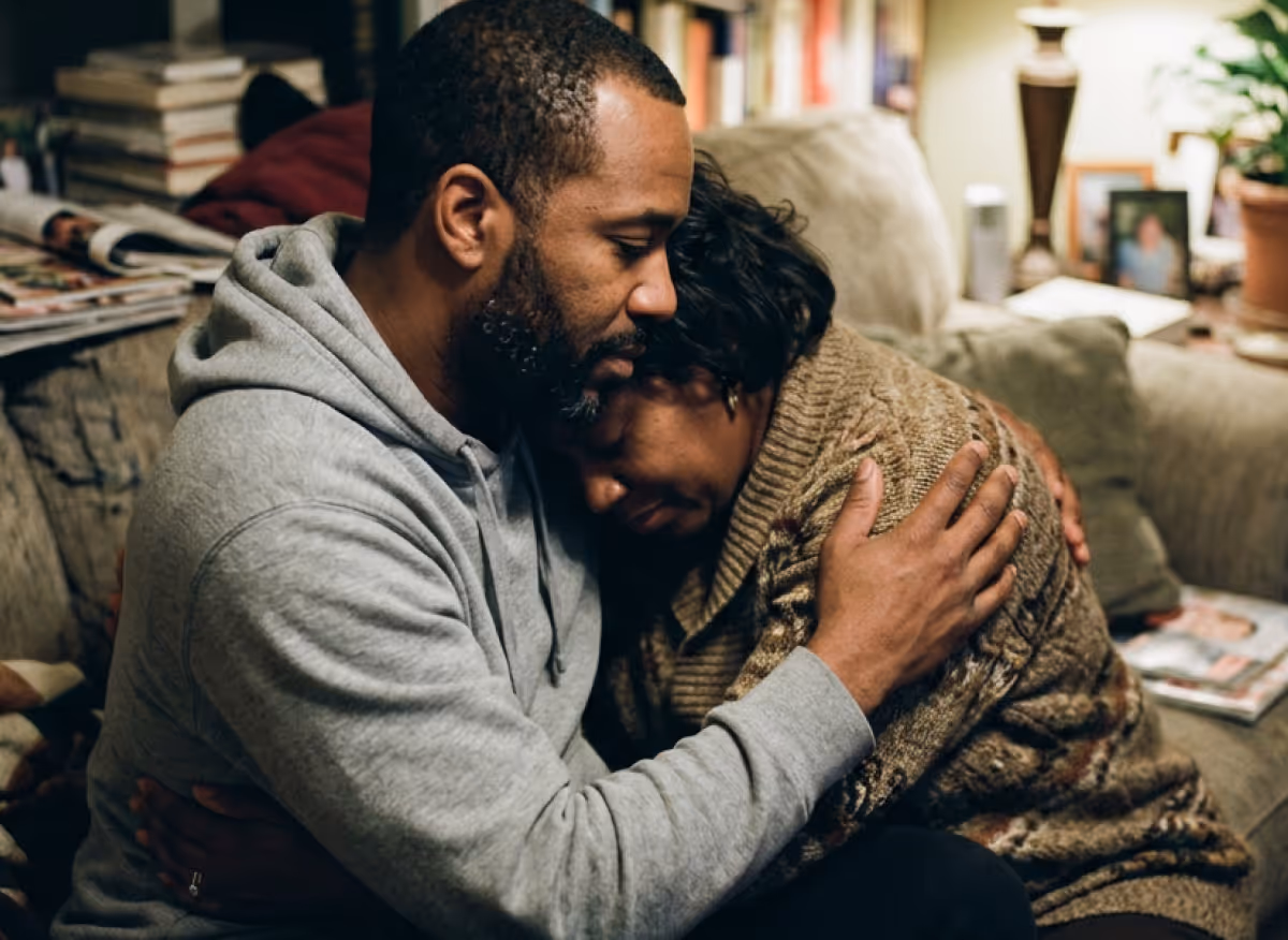 Man in gray hoodie comforting a crying woman in a brown sweater on a living room couch.