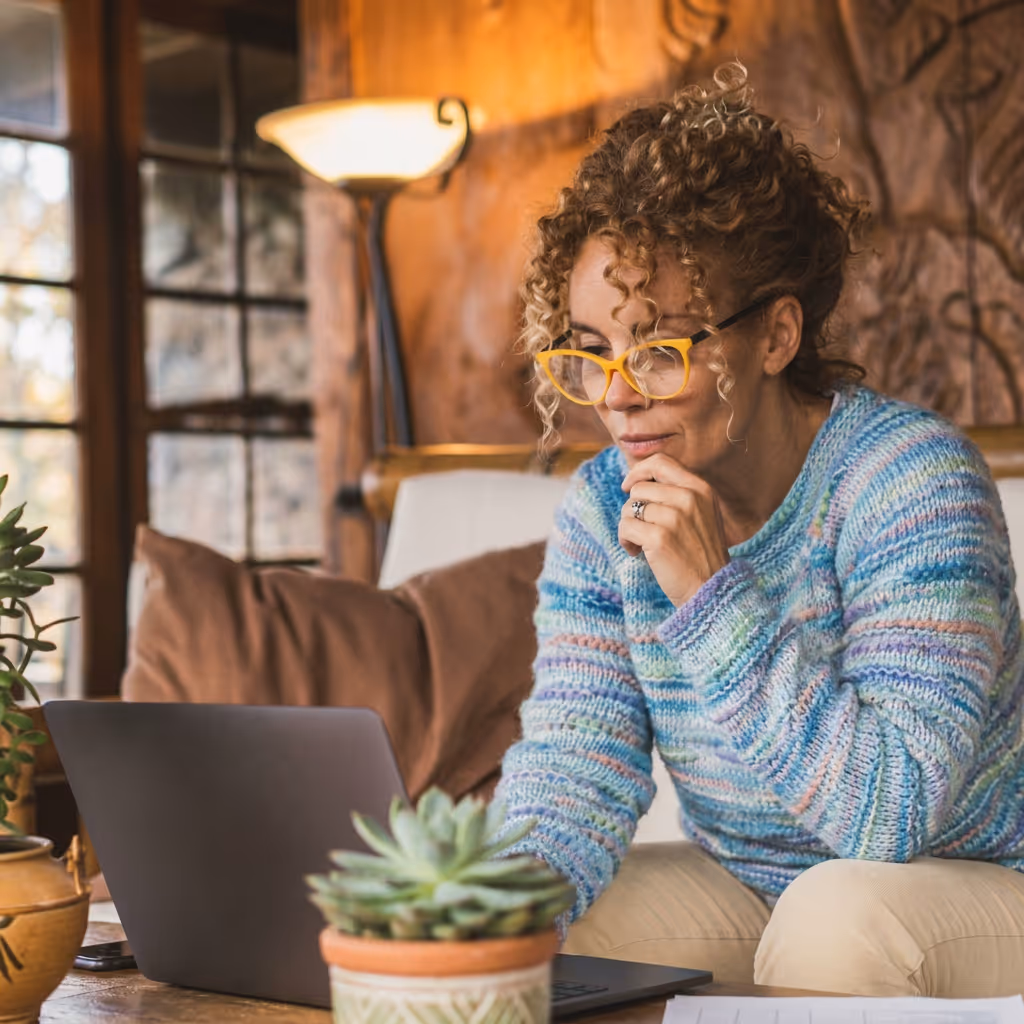 Curly-haired woman wearing yellow glasses works on a laptop in a cozy room with plants and warm lighting.