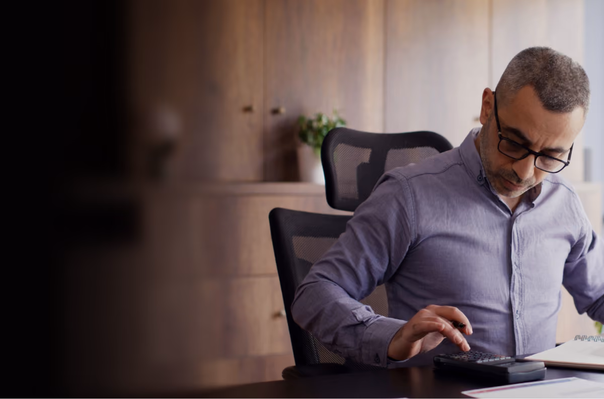 Man wearing glasses working with a calculator at a desk in an office setting.