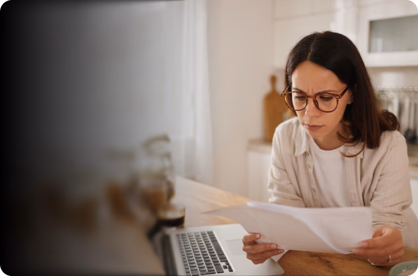 Woman with glasses looking worriedly at documents in a bright kitchen with a laptop nearby.