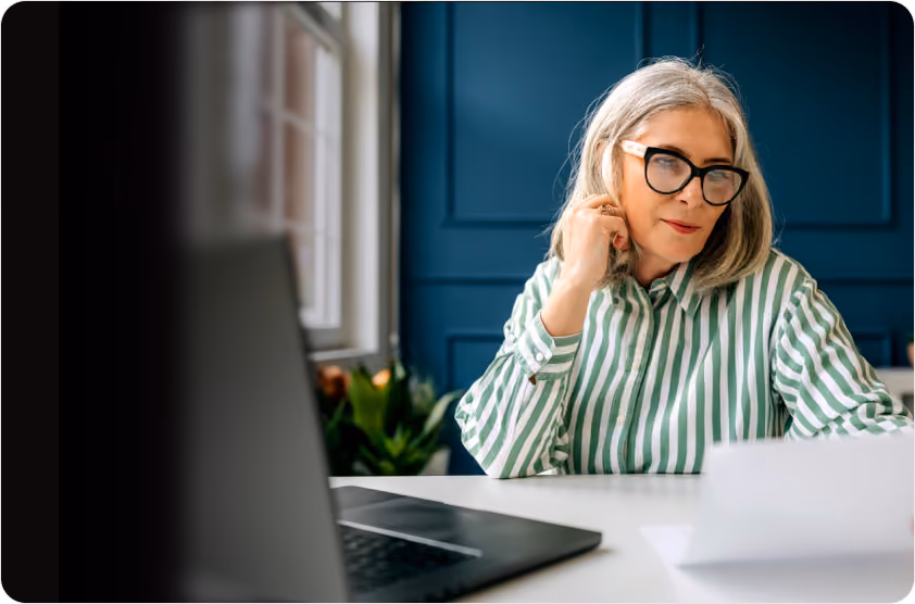 Older woman wearing glasses reviews documents at a desk with a laptop in a blue room.