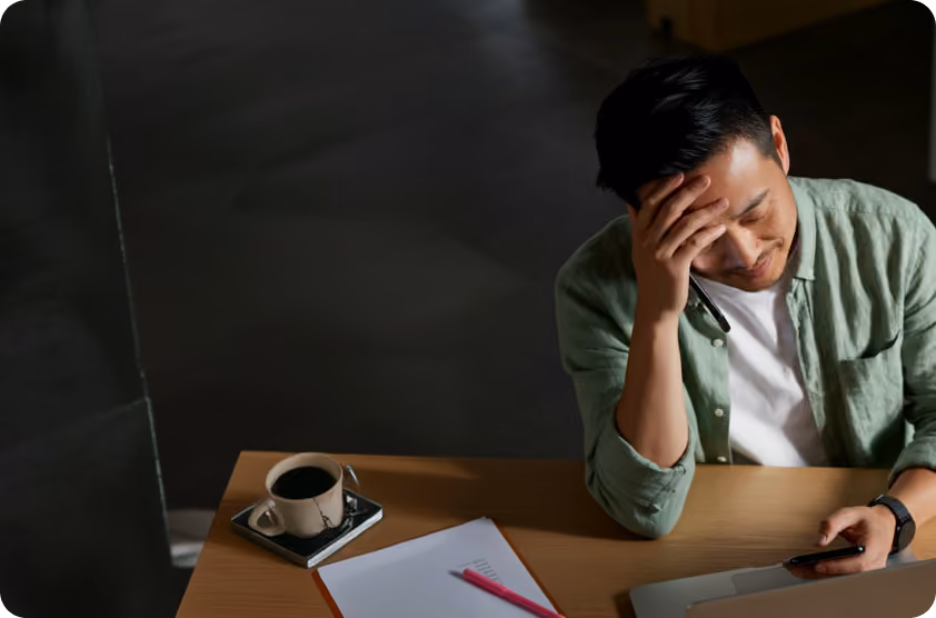 Man with stressed expression sits at desk with laptop, phone, papers, and coffee cup.