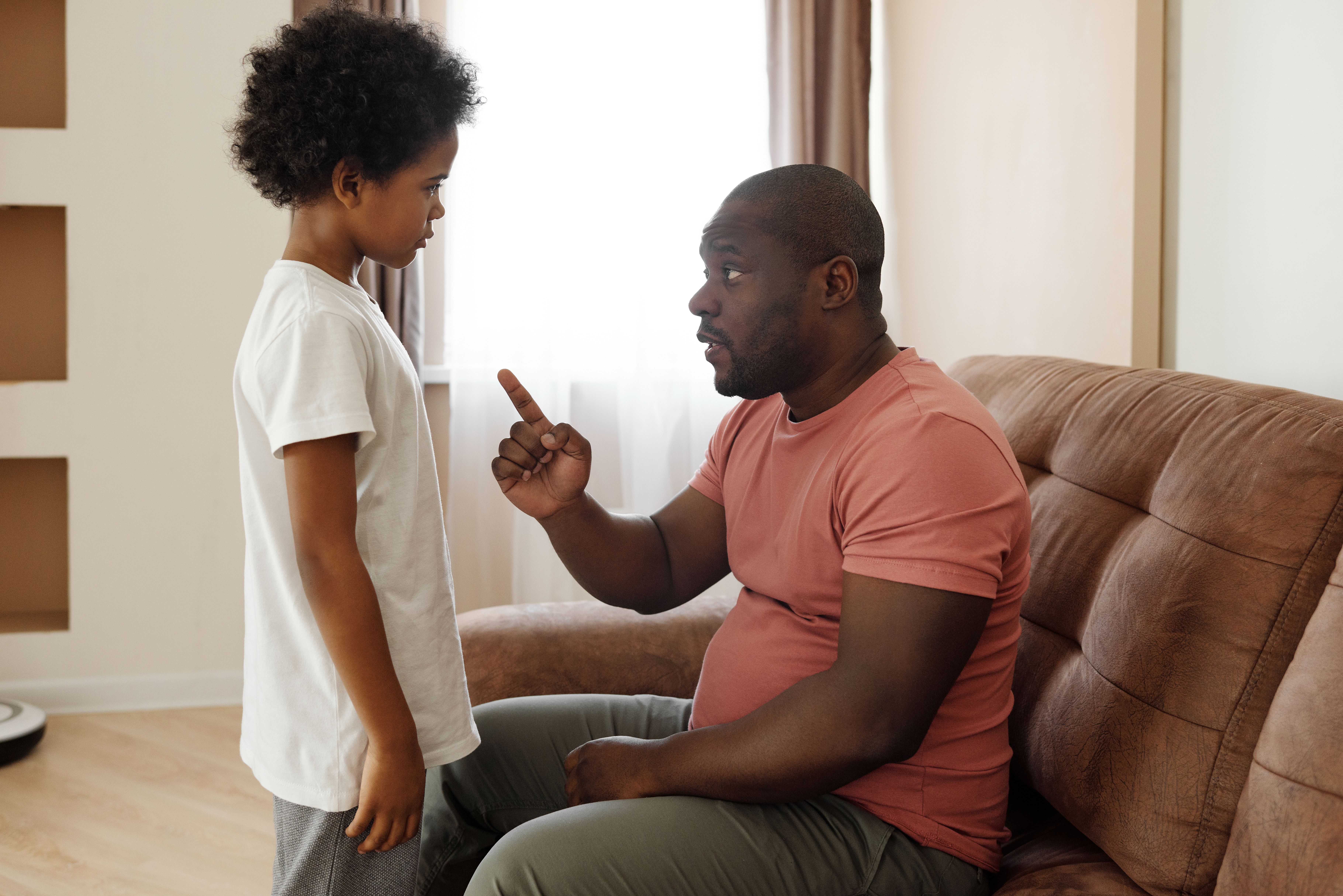 Man seated on sofa talking seriously and pointing finger at a standing boy in a living room.