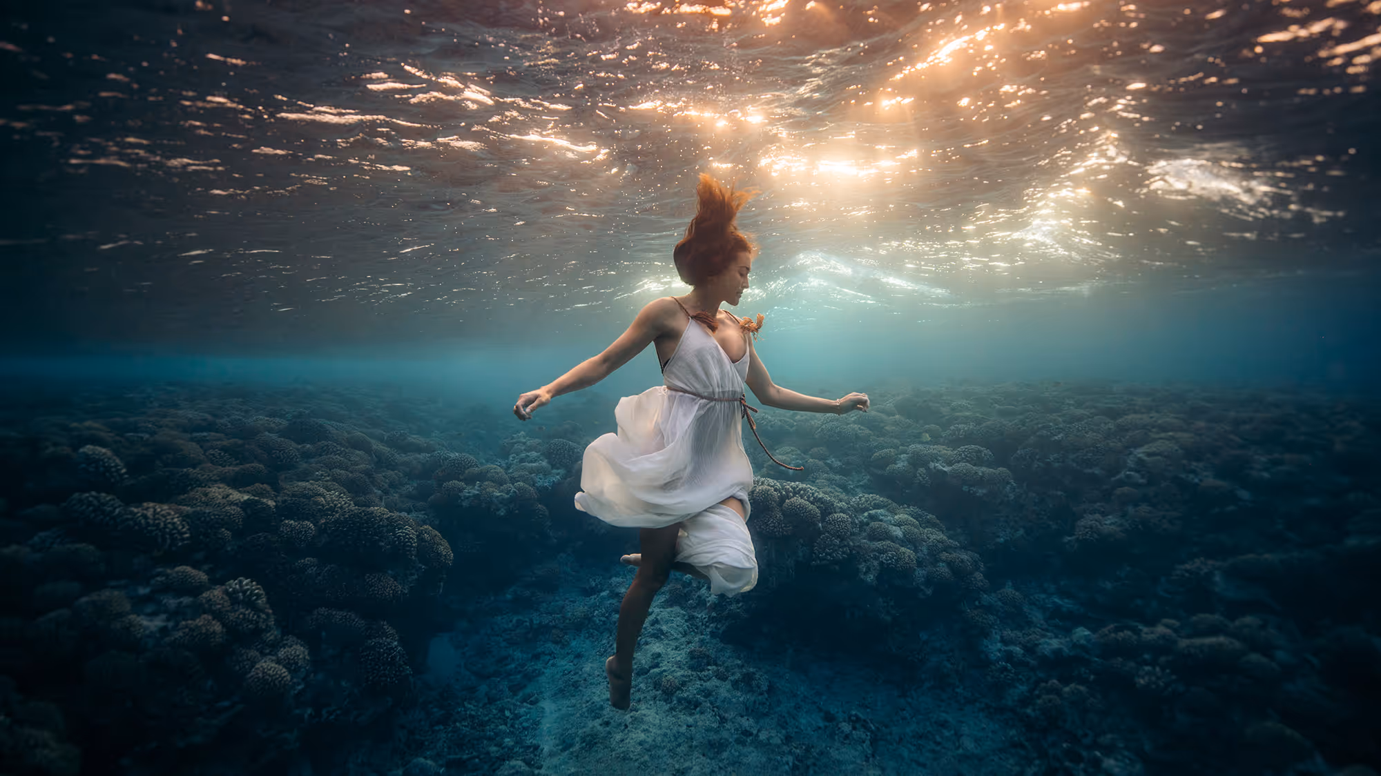 A stll frame from Emmett's short Film. Lady of the sea, of a young woman on a white dress underwater in a landscape of turquoise water and coral in French Polynesia.
