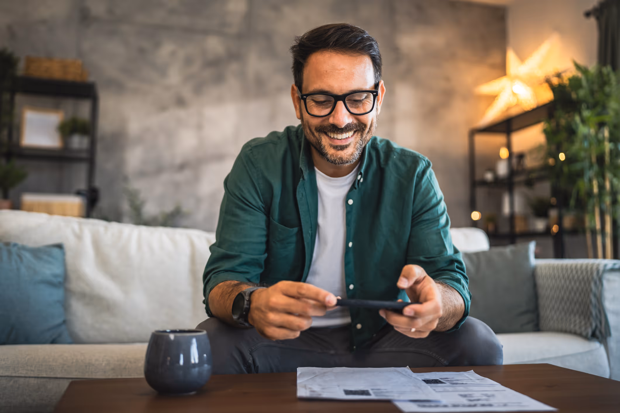 A smiling man in his late 30's in sat on a green settee. He has his payment card in one hand, his phone in the other