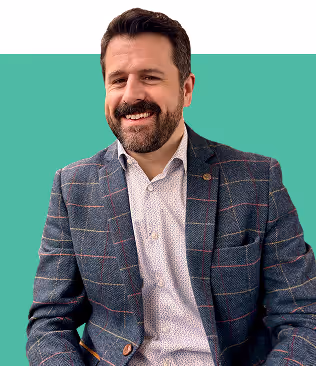 Smiling man with dark hair and beard wearing a checked blazer and white patterned shirt against a green background.