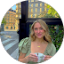 Smiling woman with long blonde hair holding a coffee cup outdoors near a green hedge.