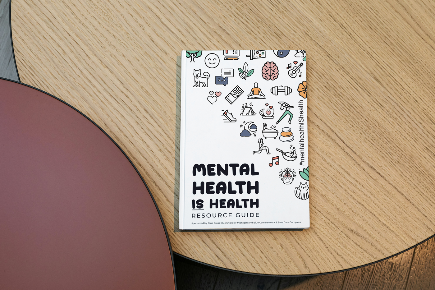A guidebook titled 'Mental Health Is Health Resource Guide' on a round wooden table with colorful mental health-themed icons on the cover.