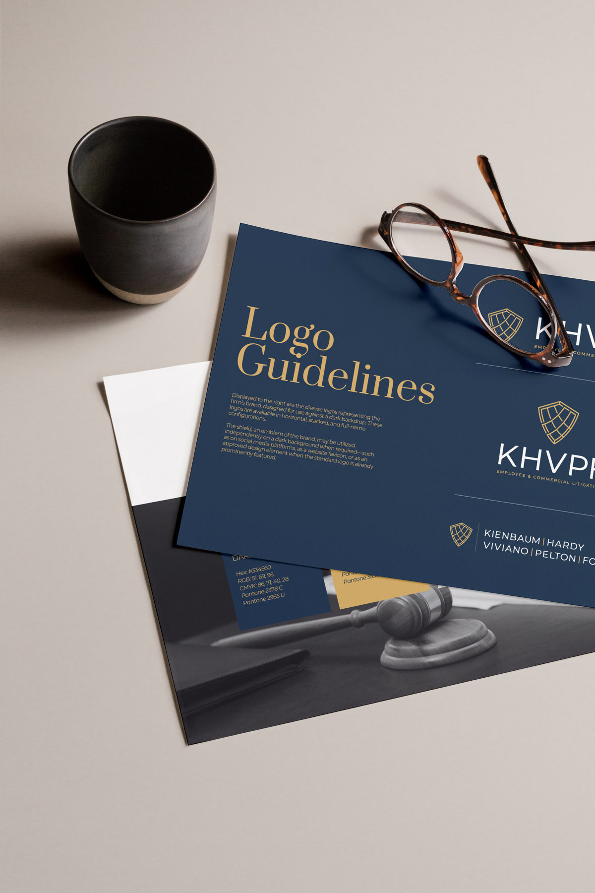 Desk scene with dark gray ceramic cup, tortoiseshell glasses, and logo guidelines sheets featuring a shield emblem and the text 'KHVP'.