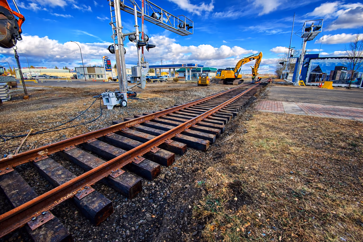 Railroad tracks under maintenance with excavator and construction workers near a railway crossing in a commercial area.