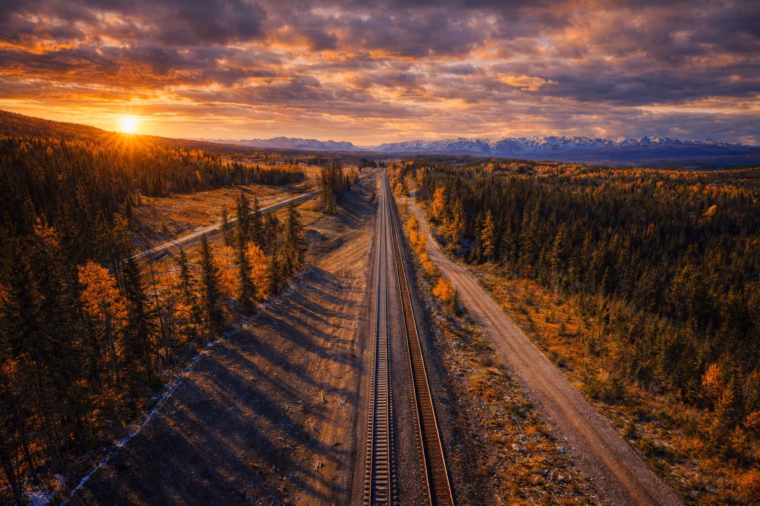 Railroad tracks running through an autumn forest at sunset with mountains in the background under a cloudy sky.