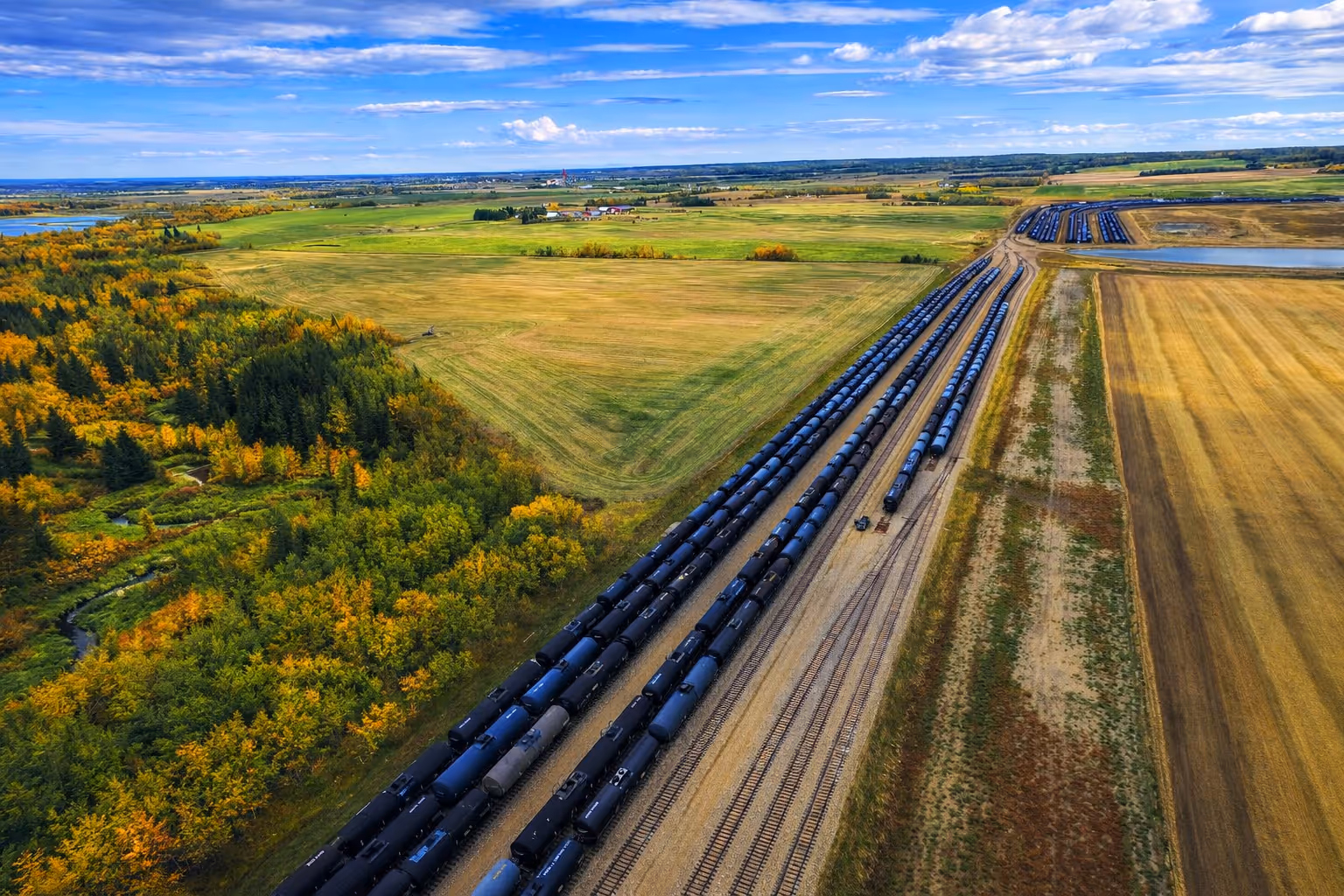 Aerial view of multiple long black tanker railcars parked on tracks next to green and yellow farming fields and a forest under a partly cloudy blue sky.