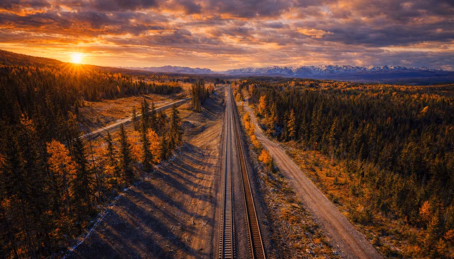 Sunset over autumn forest with railroad tracks running straight through and mountains in the distance.