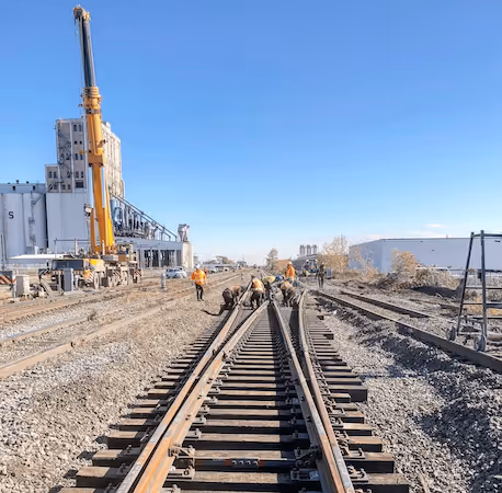 Workers performing maintenance on railroad tracks under a clear blue sky near industrial buildings and a crane.