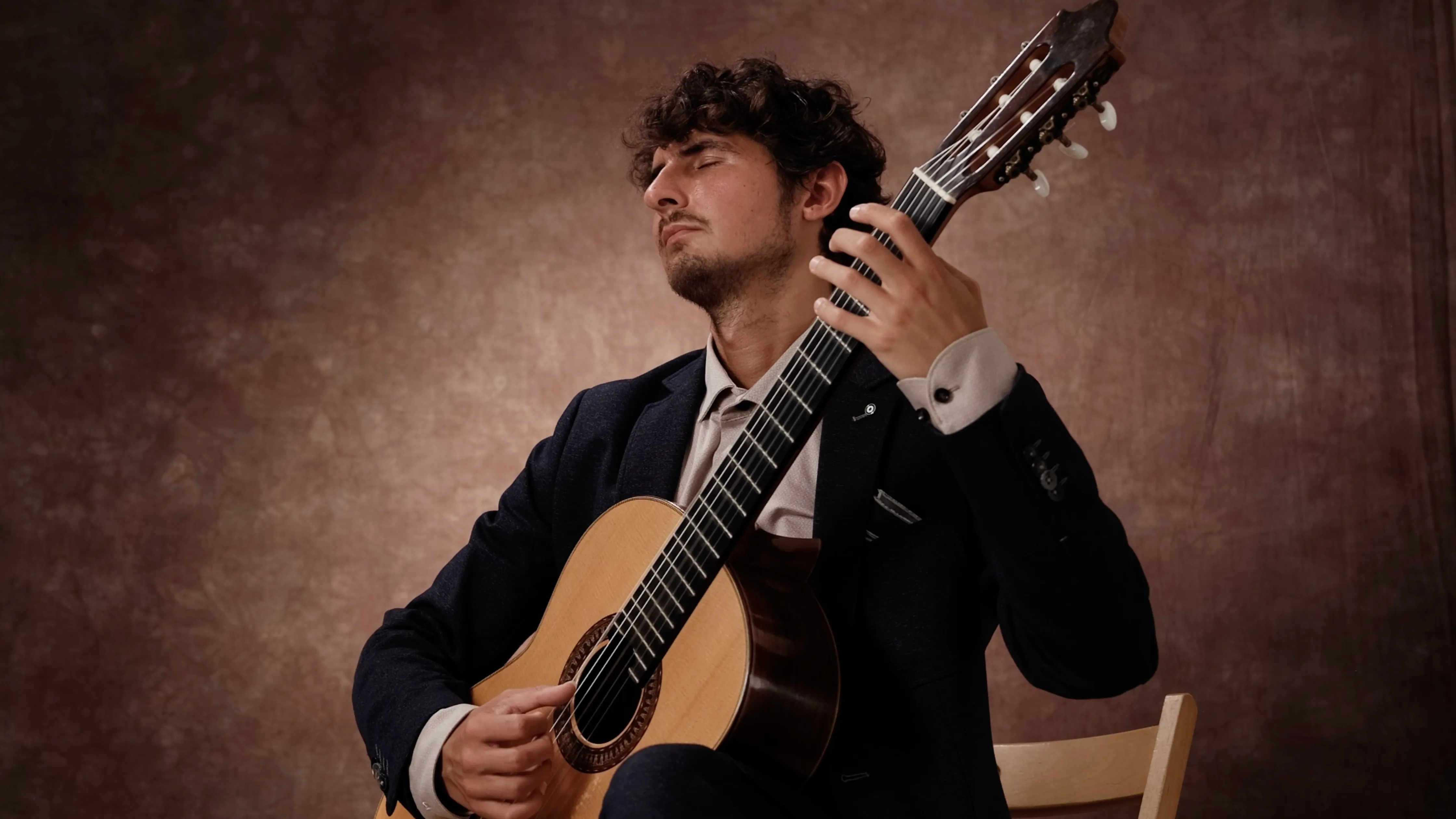 Man with closed eyes playing classical guitar seated on a wooden chair against brown backdrop.