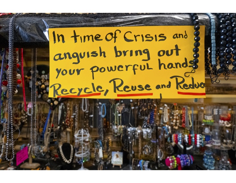 Yellow sign with black handwritten text reading 'In time of Crisis and anguish bring out your powerful hands Recycle, Reuse and Reduce' hanging above a display of various jewelry pieces.