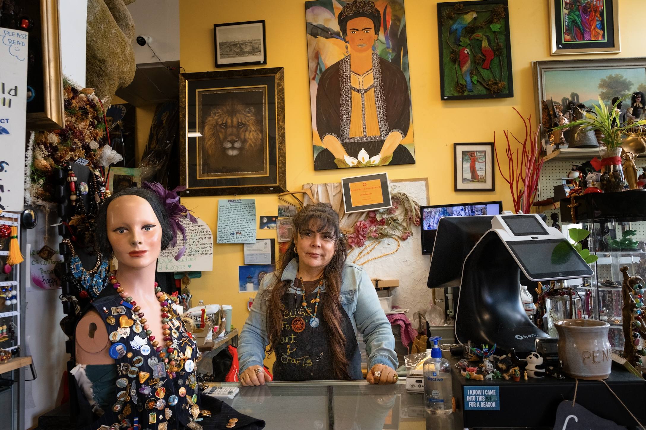 Woman standing behind a glass counter in a colorful store filled with artwork and a mannequin adorned with numerous pins and necklaces.