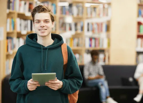 A young man holds a tablet, amidst rows of books behind him