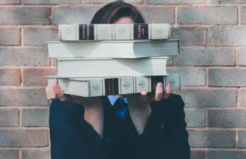 A woman holds a stack of books that cover her face.