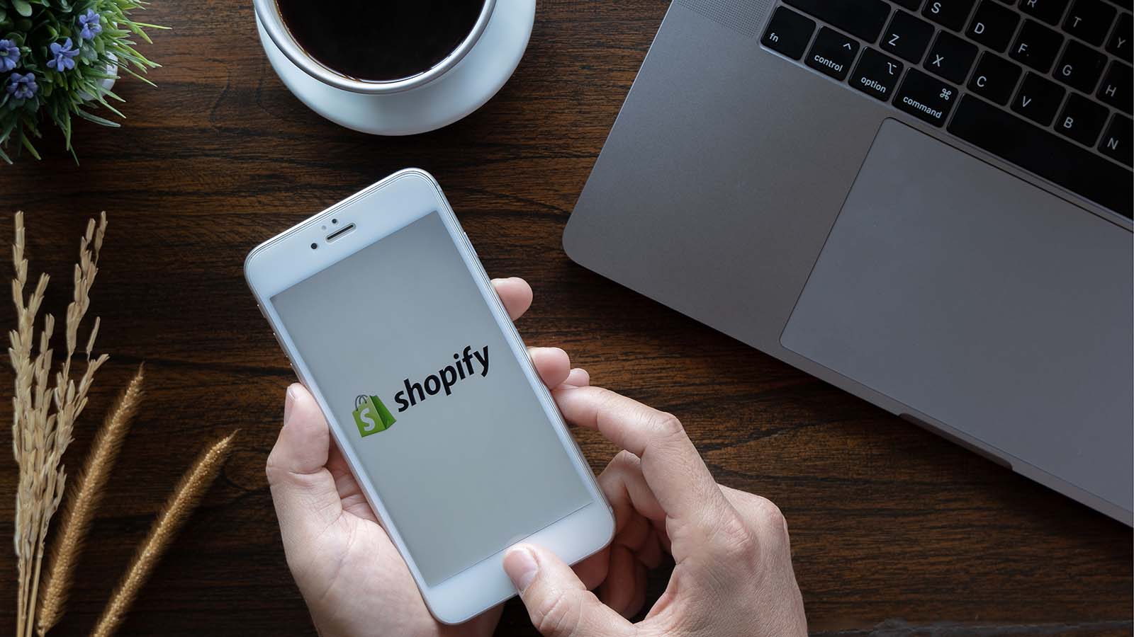 Shopify logosu görünen bir telefon ile arka planda dizüstü bilgisayar ve kahve fincanı bulunan bir stok görüntüsü