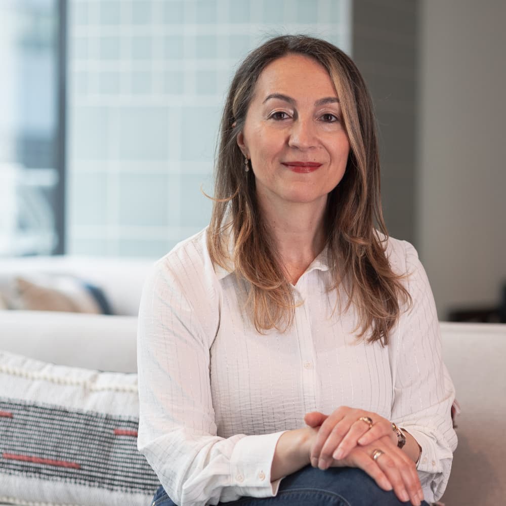 Woman with light brown hair wearing a white blouse sitting on a sofa with folded hands, smiling gently.