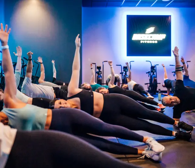 Group fitness class in a gym where participants are doing a side plank exercise with one arm raised.