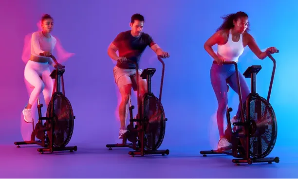 Three people exercising on stationary air bikes under colorful purple and blue lighting.