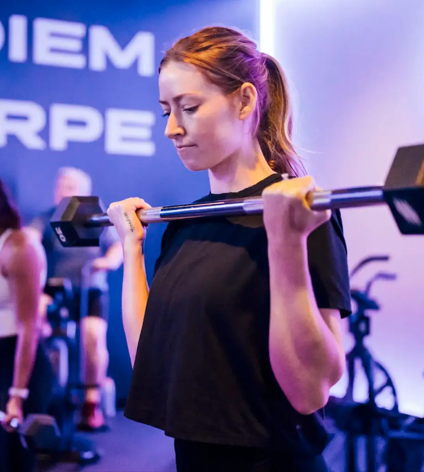 Woman wearing a black shirt lifting a barbell in a gym with purple lighting.