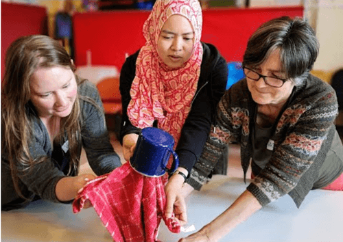 Three women are puppeteering a puppet made out of a cup and a teatowel on top of a table. One woman is young and white, one is young with light brown skin and wears a head scarf, one is an older white woman. They are all focused on the puppet.