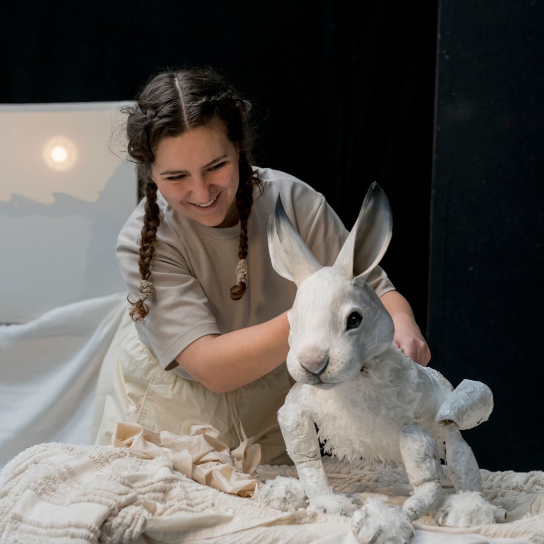 A white woman with dark hair in plaits holds a large puppet of an arctic hare, she is smiling and looking at the puppet which has an intricately carved and painted face.