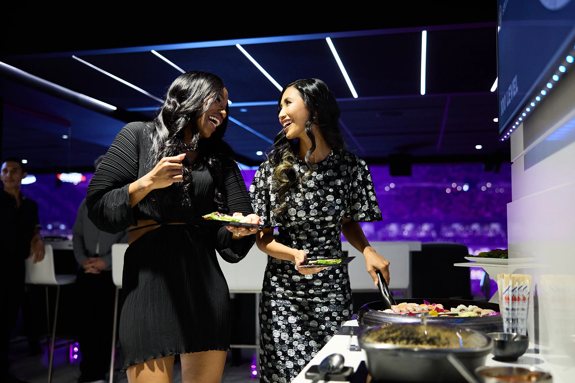 Two women smiling and chatting while serving themselves food at a buffet in a modern, dimly lit venue.