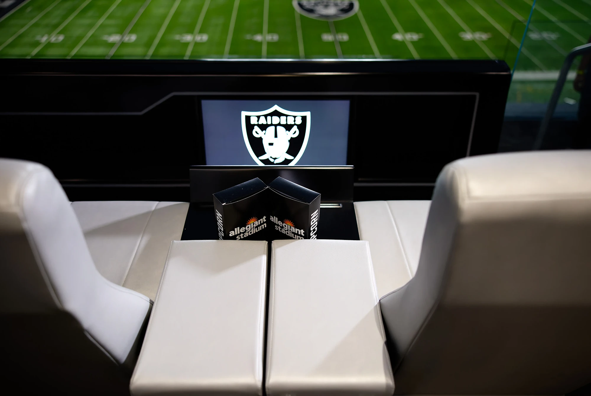 Fans seated in a luxury suite at Allegiant Stadium watching a football game, some wearing Raiders apparel.