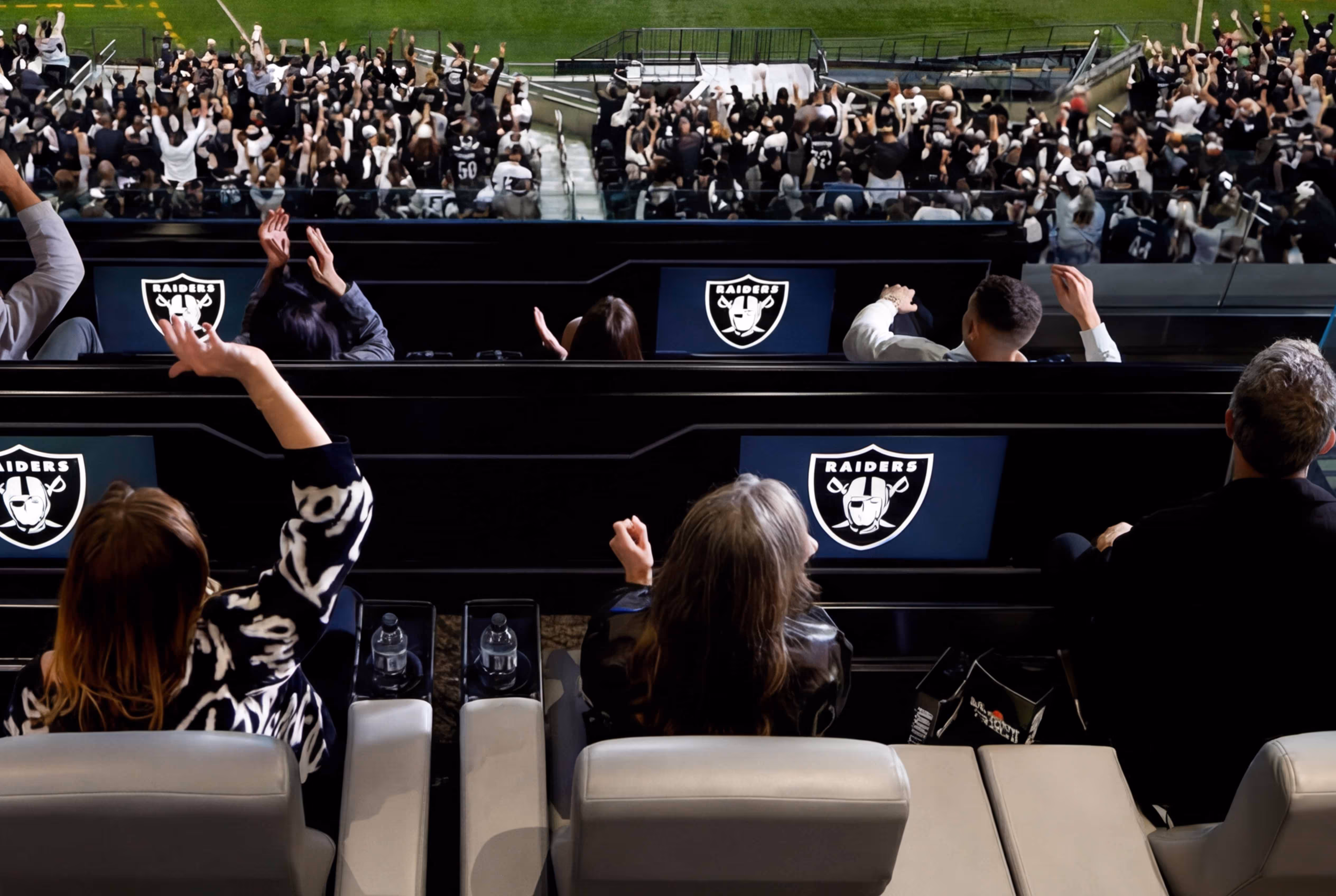 Two fans cheering in an Allegiant Stadium box watching a Raiders game on two large screens.