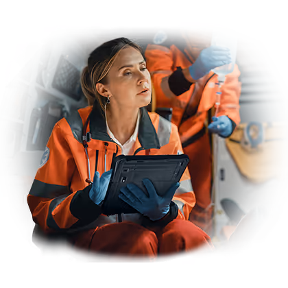 Paramedic woman wearing orange uniform and gloves holding a tablet inside an ambulance while a colleague adjusts an IV drip in the background.