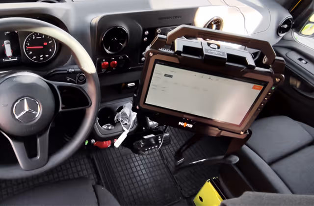 Interior of a vehicle showing a steering wheel with a Mercedes logo and a mounted rugged tablet device on a stand.