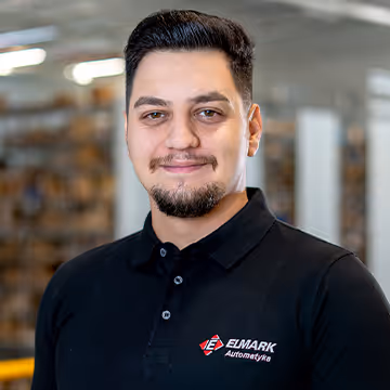 Bărbat cu păr și barbă scurtă, purtând tricou negru cu logo Elmark Automatyka, în mediu industrial.