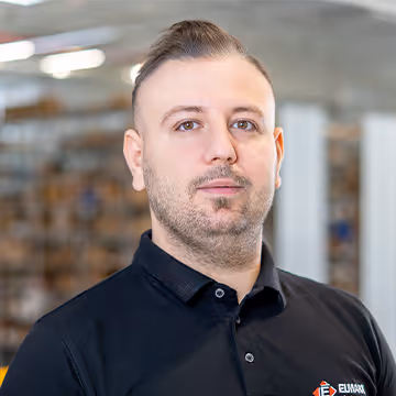 Bărbat cu păr scurt și bărbiță poartă un tricou polo negru într-un birou modern cu rafturi în fundal.