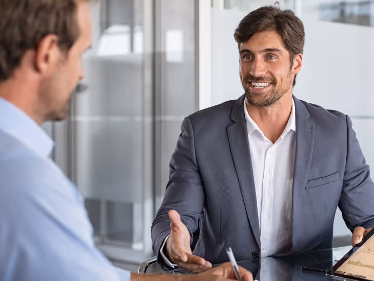 Homme souriant en costume gris clair expliquant quelque chose tout en tenant une tablette lors d'une réunion d'affaires.