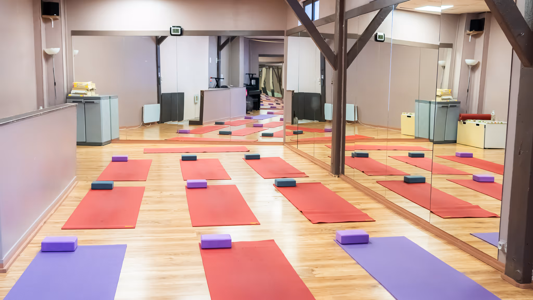 Salle de yoga avec plusieurs tapis rouges et violets alignés, chacun accompagné d'un bloc de yoga, avec un grand miroir sur un mur.